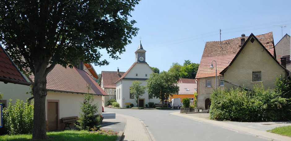 Die Straße in der Ortschaft Epplingen führt direkt auf ein Gebäude mit einem kleinen Glockenturm zu. Rechts und links sind zwei Häuser im Vordergrund. Link ist dazu ein Baum. Der Himmel ist wolkenlos blau.