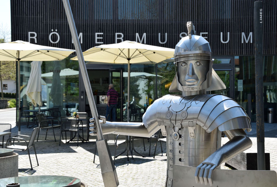 In Osterburken ist das Römermuseum, man sieht den Schriftzug oben im Bild am Gebäude. Davor sind Schirme eines Restaurants geöffnet. Am nähesten steht ein Römer in Rüstung, ein Kunstwerk aus Blech. Er hat ein Pilum, einen Schild und trägt seinen Helm.