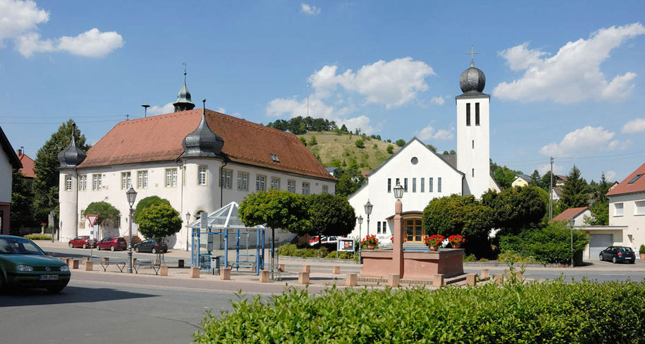 Ein romantisches Ensemble in Unterschüpf. Links ist ein Schloss, rechts eine Kirche. In der Bildmitte sind Bushaltestelle und ein Brunnen. Am nähesten ist eine grüne Hecke. Der Himmel ist blau mit weißen Wolken.