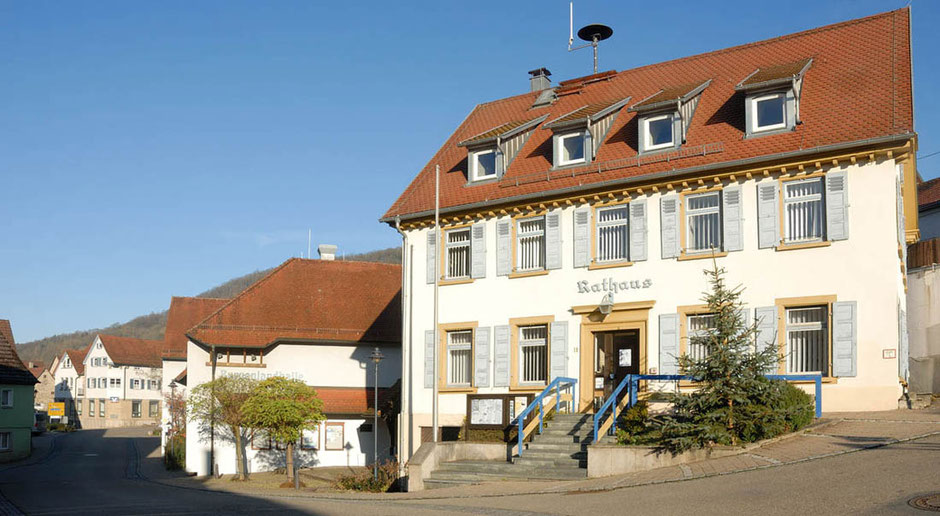 Das Rathaus von Braunsbach liegt in herrlich Sonnenschein. Der Himmel ist wolkenlos. Eine Treppe führt hinauf zum Eingang. Die Straße, die links im Bild in die Tiefe führt, liegt im Schatten.