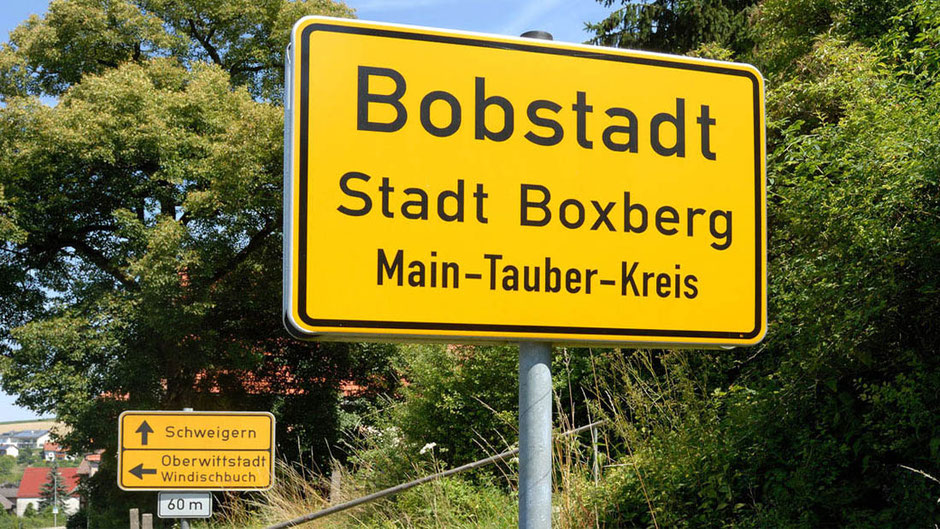Vor einer Böschung sieht man zwei Verkehrsschilder. Das nähere ist das Ortseingangsschild von Bobstadt, es ist sehr dominant. Etwas weiter hinten sieht man ein Hinweisschild zu anderen Orten. Der Hintergund ist grünes Baumlaub und Wiese.