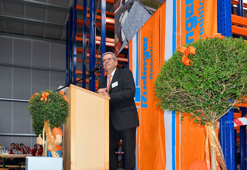 Roland Rüdinger steht mit einem schwarzen Anzug und orangener Krawatte hinter einem Rednerpult und lächelt. Vor dem Pult ist eine Pflanze, rechts im Bild ebenso. Hinter dem Redner sind zwei Rüdinger-Fahnen und Regale sichtbar.
