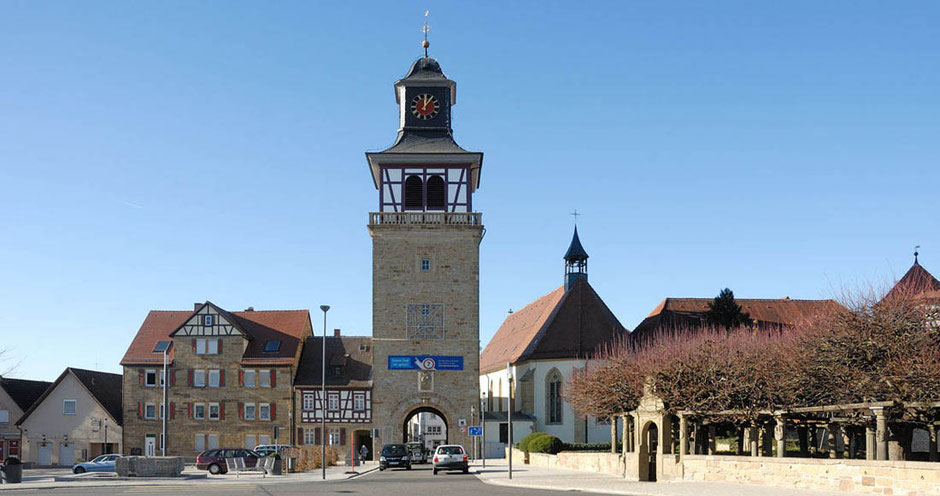 Ein absolut beeindruckend hoher Stadtturm ist dominant. Durch ihn führt die Straße vom Betrachter hin und hindruch in die Innenstadt von Neuenstadt. Links sind historische Gebäude, rechts ist eine Kirche. Vorne rechts ist eine Gartenanlage. 
