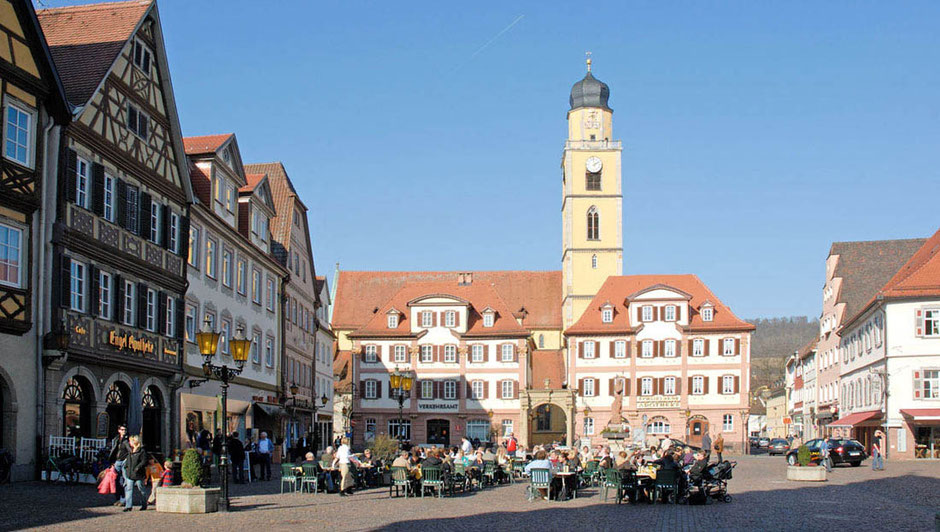 Eine "Schokoladen-Ansicht" in Bad Mergentheim. Man sieht auf zwei barocke Häuser mit einem Glockenturm. Davor ist eine große Fläche nur für Fußgänger. Fast komplett sind Tische mit vielen Menschen belegt. Die Sonne scheint auf die ganze Szenerie.