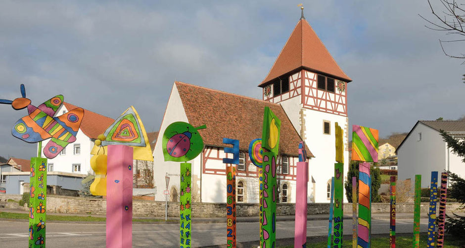 Hinter rund zehn Stelen, alle herrlich bunt, von Kindern gemalt, steht die kleine Kirche in Weißach. Es ist ein Fachwerkbau. Zwischen Kunstwerken und Kirche führt eine Straße, rechts und links sind aktuelle Wohnhäuser.