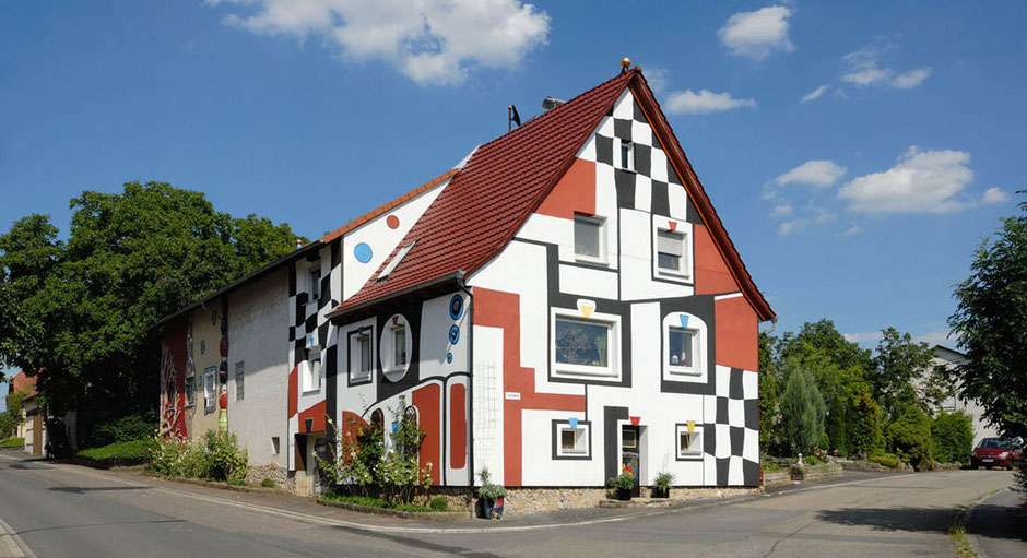 Das bekannteste Wohnhaus in Schwabhausen. Im Hundertwasser-Stil ist es eine Mischung aus roten Flächen und Schachbrett-Muster. Davor gabelt sich die Straße. Rund um das Haus ist viel Grün. Der Himmel ist blau, das Haus ist in der Sonne.