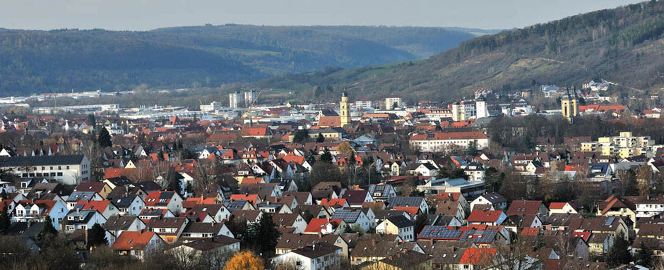 Man blickt von einer Anhöhe auf ganz Lauda-Königshofen. Im Hintergrund ist Wald im Dunst zu erkennen. In der Bildmitte sieht man eine Kirche, am rechten Bildrand noch eine.