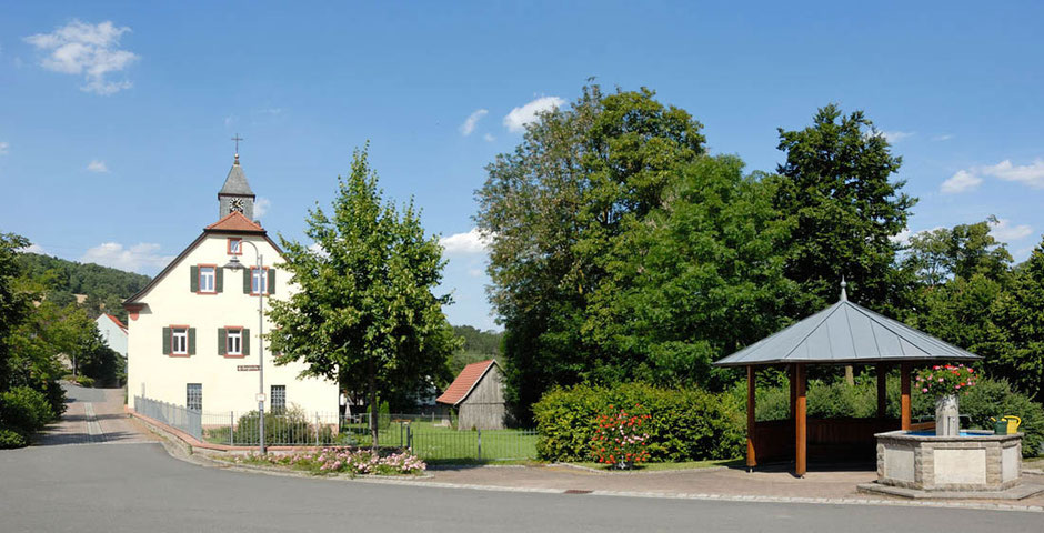 Ein bekanntes Motiv in Angeltürn. Eine Straße führt an einem Gebäude mit einem kleinen Glockenturm auf der Mitte des Daches. Vorne rechts ist ein hölzernes offenes Häuschen neben einem kleinen Brunnen.