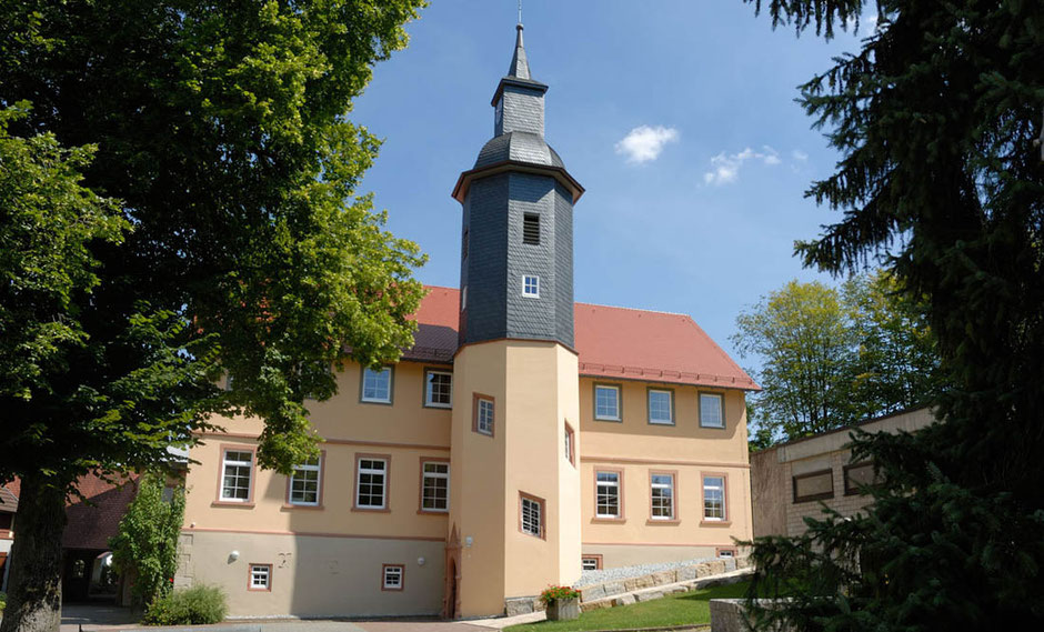 In Oberschüpf sieht man auf ein zweistöckiges Wohnhaus in Beige, mit einem Turm an der Vorderseite. Rechts und links und auch im Hintergrund stehen Bäume. Die Himmel ist blau und fast wolkenlos, das Haus liegt in der Sonne.