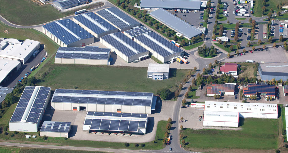 Auf etwa zwei Dritteln der Luftaufnahme sieht man 13 Rüdinger-Lagerhallen, alle mit Fotovoltaik. Links knapp und rechts sind andere Firmengebäude sichtbar. Rechts sieht man auch eine Straße, eine Wendeplatte und einen Parkplatz.