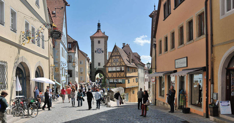 Die bekannteste Ansicht von Rothenburg ob der Tauer. Ein Stadttor ist in der Mitte. Rechts und links flankieren historische Gebäude die Straße dorthin. Viele Touristen sind unterwegs. Vor dem Turm steht ein berühmtes Fachwerkhaus. Der Himmel ist blau.