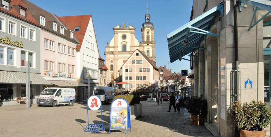 Die Fußgängerzone in Neckarsulm bei herrlichem Sonnenschein und blauem Himmel. Rechts und links sind Geschäfte in Wohnhäusern. Man blickt auf die Kirche zu, davor steht ein weiteres Wohnhaus. Es ist eine historische Kirche.