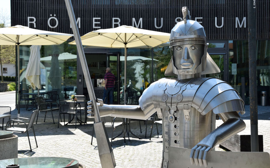 Ein Kunstwerk aus Blech dominiert vorne das Bild. Es ist ein Römer mit einem Pilum und seinem Schild und Helm. Er steht vor einem Außen-Restaurant-Bereich. Der wiederrum befindet sich vor dem Römermusem: Man kann das Wort am oberen Bildrand lesen.
