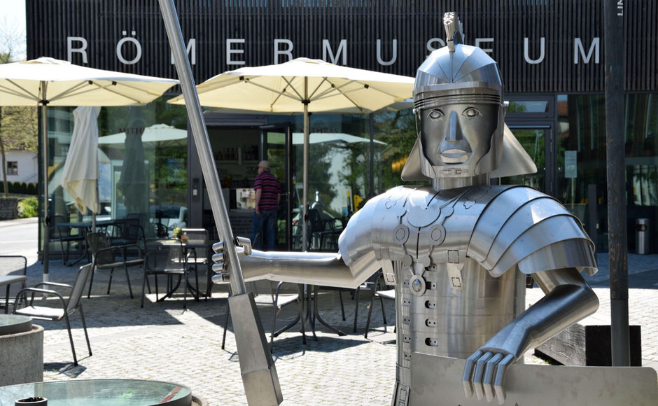 Vor dem Römermuseum, dessen Schriftzug man am Gebäude lesen kann, steht, als Kunstwerk, eine Blech-Skulptur eines Römers. Man sieht ihn bis zur Bauchhöhe. Er hält ein Pilum, hat einen Helm auf und hat in der anderen Hand seinen Schild. 
