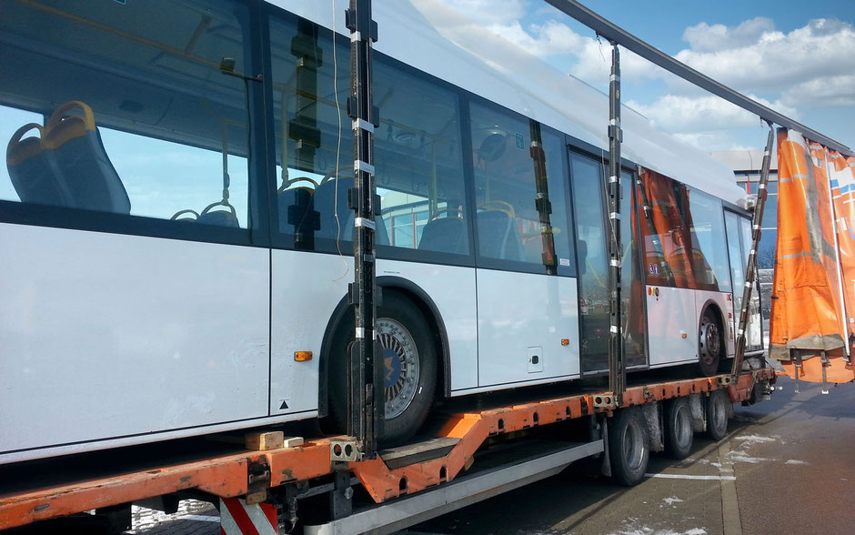 Man sieht einen komplett geöffneten Anhänger eines Planentiefladers von schräg vorne. Das Fahrerhaus ist nicht zu sehen. Auf der Ladefläche ist ein weißer Linienbus. Rechts sieht man die offene Plane.