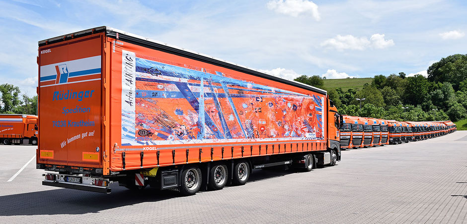 Ein großer Lkw steht vor einer Reihe geparkter Lkw. Auf der Lkw-Plane sieht man ein selbstgestaltetes abstraktes Kunstwerk in den Farbtönen orange blau und weiß.  