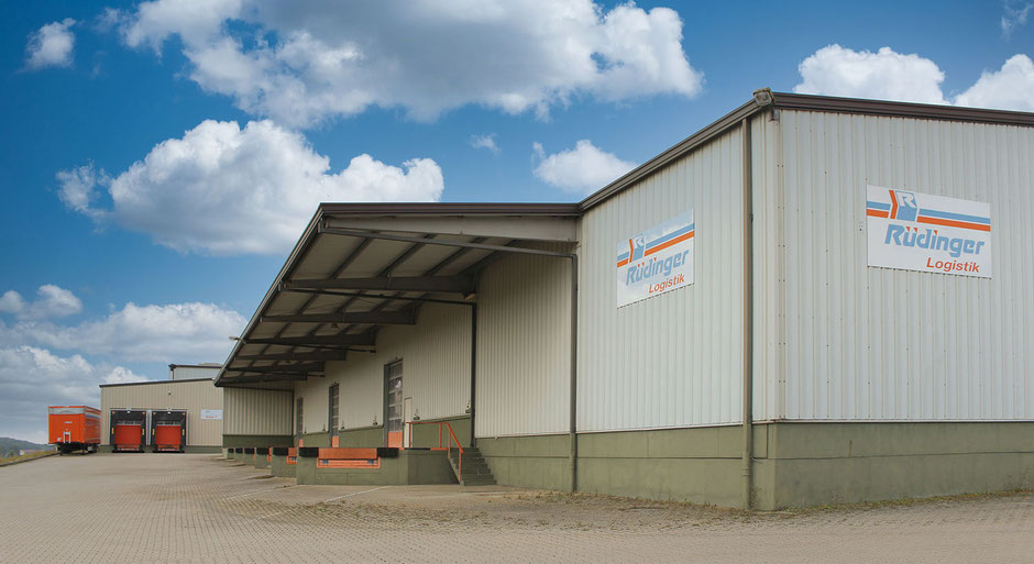 Man sieht die erste 1995 entstandene Lagerhalle in Boxberg. Man blickt seitlich an der Außenfassade entlang. Zwei Schilder weisen auf die Spedition hin, im Hintergrund sieht man drei Anhänger Speditions. Der Himmel ist blau mit vielen weißen Wolken.