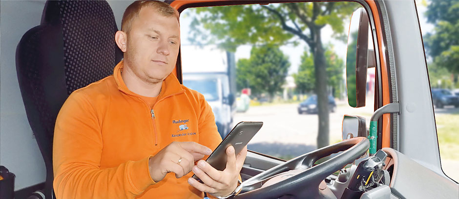 Ein Lkw-Fahrer mit orangenem Sweatshirt sitzt in der Fahrerkabine seines Lkw. Er hält ein Tablett in der Hand und tippt mit der anderen Hand etwas ein.