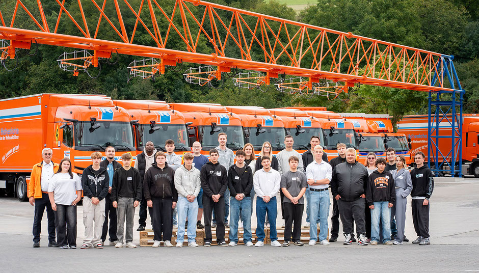 26 Auszubildende stehen vor acht E-Lkw. Die E-Lkw stehen unter dem E-Lkw-Ladepark, einem Gerüst, das wie eine Brücke über die Lkw führt.ist.
