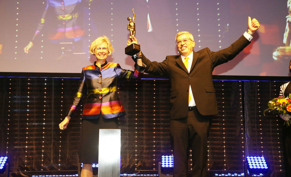 Anja und Roland Rüdinger sind auf der Bühne und sichtbar stolz auf den Erhalt einer goldenen Auszeichnung. Sie heben diese Auszeichnung gemeinsam in die Höhe. Roland Rüdinger zeigt noch mit dem Daumen nach oben am gestreckten Arm.