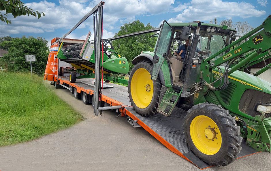 Auf einem Weg in grünem Umfeld sieht man schräg von hinten auf einen komplett geöffneten Planentieflader. Das Fahrerhaus ist nicht zu sehen. Auf der Ladefläche steht ein landwirtschftliches Gerät. Auf der hinteren Ladebordwand steht ein Traktor.