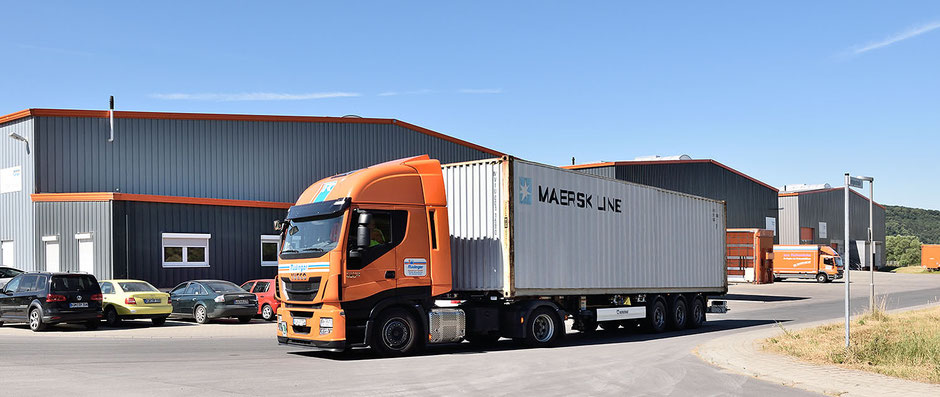 Ein Planentieflader mit Chassis und Container verlässt das Betriebsgelände. Im Hintergrund sind drei neu erbaute Lagerhallen und man sieht zwei weitere Lkw. Dazu vorne auch noch vier Pkw.