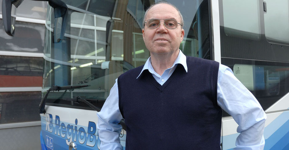 Ein Busfahrer mit blauem Hemd und dunkelblauem Polunder steht vor einem Regio-Bus, von dem man fast nur die Frontscheibe erkennt. Der Bus selbst steht vor einer Wartungshalle, von der man das Tor sieht.