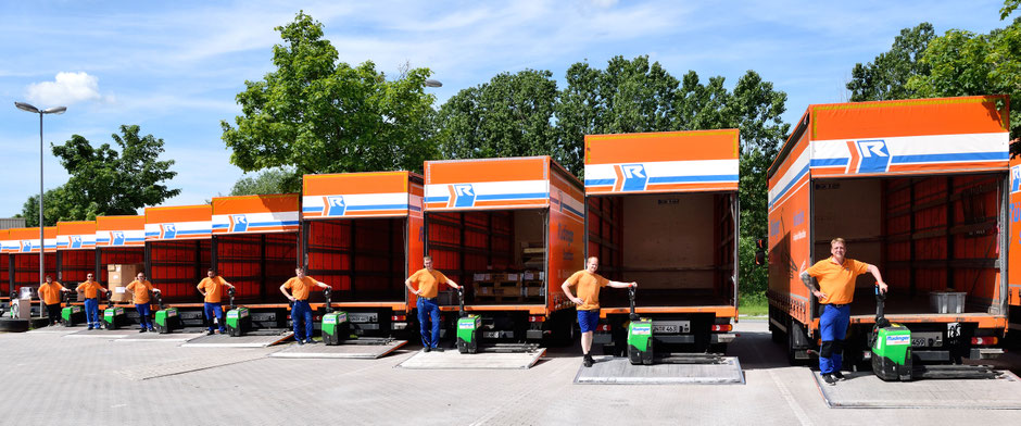 Man sieht eine Aufstellung von neun 7,5-Tonnen-Lkw von der Rückseite. Alle Ladeklappen sind heruntergelassen. Es stehen acht Mitarbeiter mit Kleidung in den Farben der Spedition und stützen sich jeweils auf eine Elektro-Ameise. 