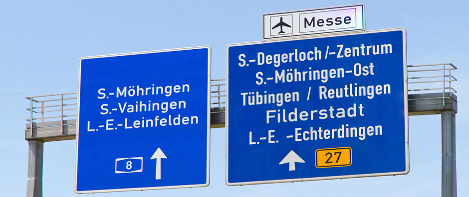 Die Schilderbrücke über einer Autobahn. Weiße Schrift auf Dunkelblau. Es sind zwei Tafeln. Unter anderem geht es zum Flughafen Stuttgart.