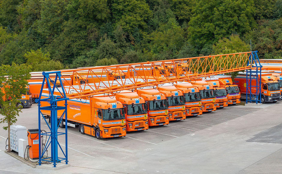 Acht Rüdinger-E-Lkw stehen unter der einer Ladeeinrichtung. Diese Ladeeinrichtung ist in orange und blau gehalten. Rechts daneben stehen weitere Lkw.