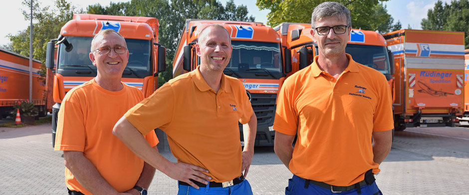 Drei Rüdinger-Mitarbeiter lachen zur Kamera. Alle tragen orangene Shirts. Im Hintergrund sieht man fünf Lkw.