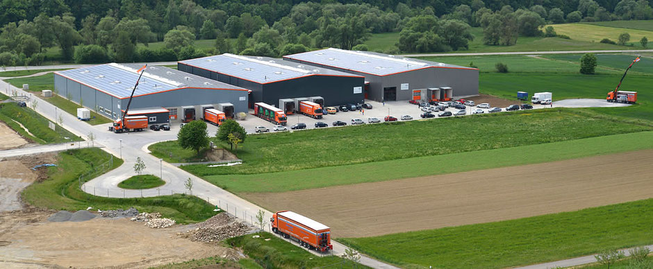 Es ist ein Foto in Dörzbach von einem nahen Weinberg, fast eine Luftaufnahme. Zwischen Wald und Grünflächen rechts und unten sind drei neue Lagerhallen. Links ist eine Straße mit Wendeschleife. Mehrere orangene Lkw sind sichtbar.