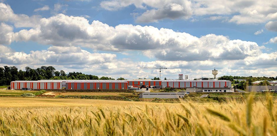 Man schaut über ein reifes Gerstenfeld im kompletten unteren Bilddrittel. Darüber ist ein waagrechter Bereich mit vier neuen Lagerhallen, dahinter ist links Wald und die obere Hälfte ist blauer Himmel und viele weiße Wolken.