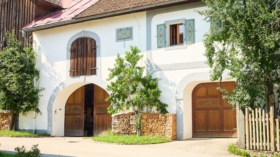 Auf der Straßenseite des Biohof Stadlers sind zwei schöne Holztore zu sehen. Die Fenster haben Fensterläden mit Rautenmuster und ein Holzstoß mit Holz für den Küchenofen ist zwischen den Toren Vor dem Holzstoß steht ein grüner Nektarinenbaum.