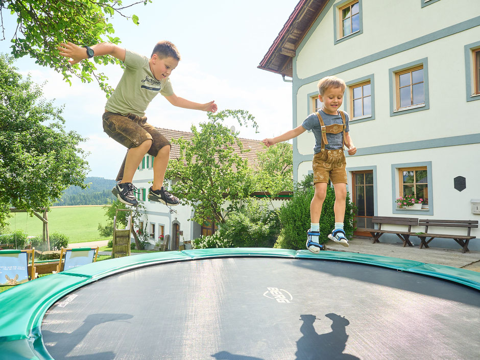 Zwei Buben in kurzen Lederhosen springen fröhlich auf dem großen Gartentrampolin. Im Hintergrund sieht man den malerischen Bauernhof.