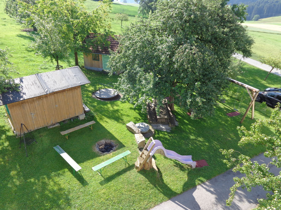 Aussicht in den Garten mit Feuerstelle, die von Sitzbänken umrahmt ist, Wellenrutsche, Karussell, Schaukel, großer Esstisch unterm Apfelbaum, Gartenhütte, Bienenhütte und Parkplatz