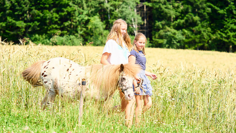 Zwei Mädchen in kurzen Kleidern führen das weiße Pony entlang eines Getreidefeldes.