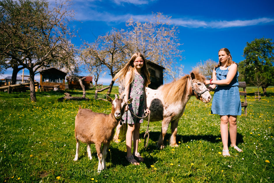Im Hintergrund sind ein strahlender Himmel und blühende Bäume, Spielgeräte und zwei Holzhütten zu sehen. Groß im Vordergrund sind zwei fröhliche Mädchen mit einer Ziege und einem Pony, die die Tiere halten.