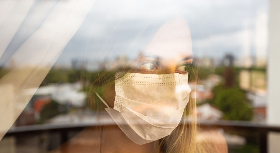 Frau mit Mund-Nasen-Schutz-Maske steht am Fenster eines Hotels und schaut traurig raus, weil sie wegen Corona in Quarantäne ist. Der Ergänzungs-Schutz Covid-19 zahlt die Hotelkosten