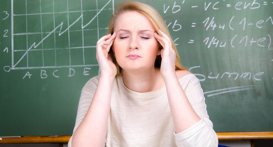 Eine Lehrerin mit Kopfschmerzen hält sich den Kopf vor einer Schultafel