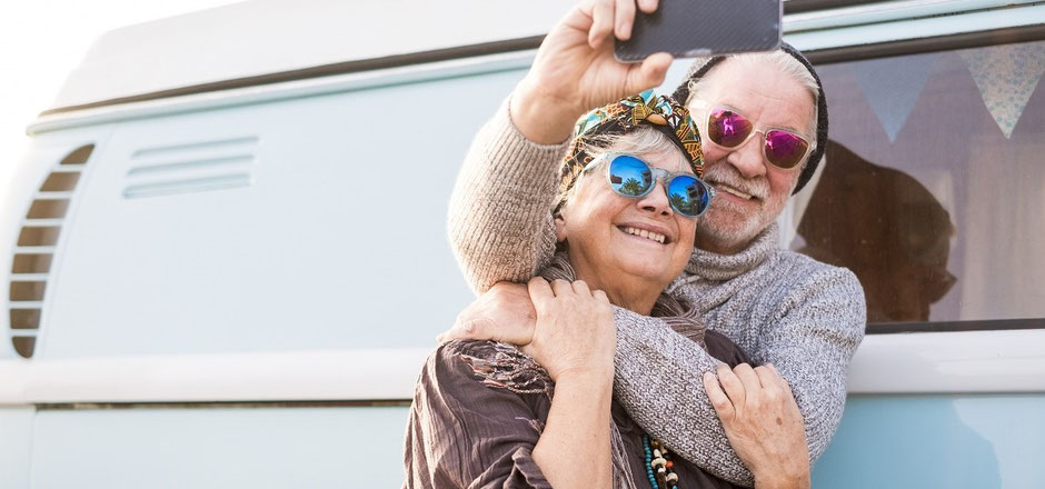 Älteres Ehepaar steht im Urlaub vor einem Campingbus und macht Selfies. Gut geschützt mit einer Jahres-Reiseversicherung.