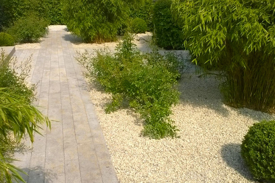 Das Bild zeigt einen Gartenweg aus hellen Steinplatten, der sich durch einen mit hellem Kies belegten Garten schlängelt. An den Seiten des Weges wachsen diverse Sträucher und Bambuspflanzen. Der Gesamteindruck ist ruhig, modern und minimalistisch.