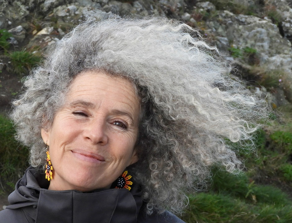 Vicki Cooper, Künstlerin und Körpertherapeutin mit grau-gelocktem im Wind wehendem Haar, draussen vor einem Felsen, lächelt ruhig in die Kamera. Sie trägt farbige Ohrringe und eine dunkle Jacke.