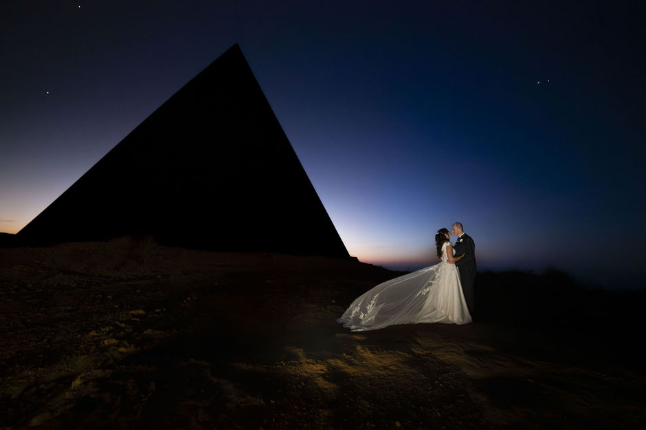 Sposi in un abbraccio romantico durante il tramonto in Sicilia, con una maestosa silhouette architettonica sullo sfondo. Fotografia di matrimonio artistica e scenografica a cura di Daniele Benenati, fotografo a Palermo.
