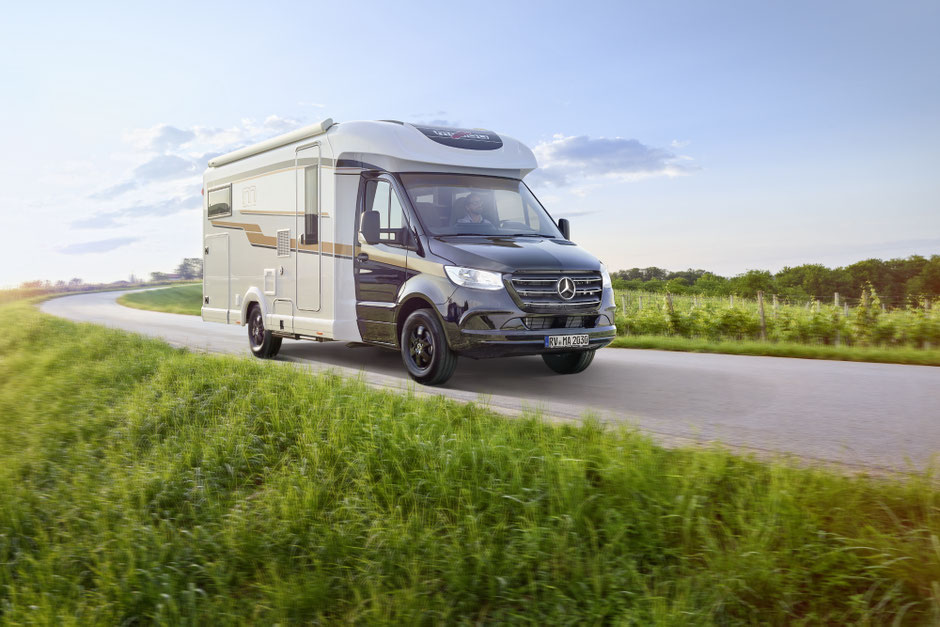 Ein modernes teilintegriertes Wohnmobil auf Basis eines schwarzen Mercedes-Benz Sprinter fährt auf einer schmalen Landstraße durch eine grüne Wiesenlandschaft unter blauem Himmel