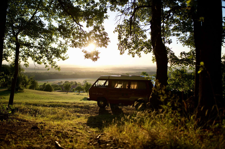 Ein dunkler Campingbus steht im Schatten großer Bäume auf einer Wiese, während die tiefstehende Sonne durch das Laub bricht und eine weite Hügellandschaft im Hintergrund beleuchtet. 