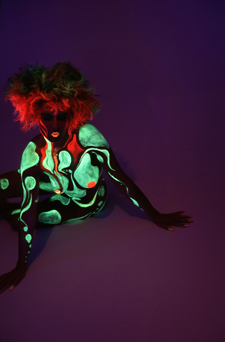 A nude woman with fluorescent make up is sitting on the studio floor