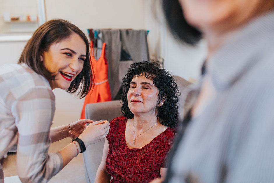 Woman in a red top with curly hair having her make up done whilst her female friend is laughing at the camera