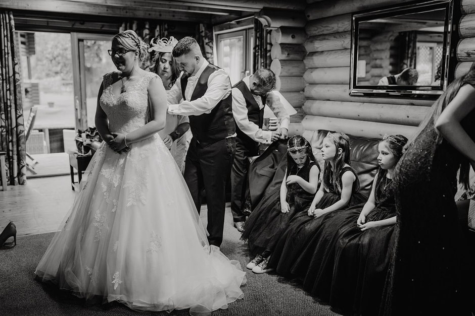 black and white photo of a bride being fastened into her dress whilst three flower girls look on