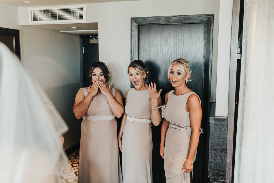 three bridesmaids with happy and shocked faces when seeing the bride in her dress for the first time